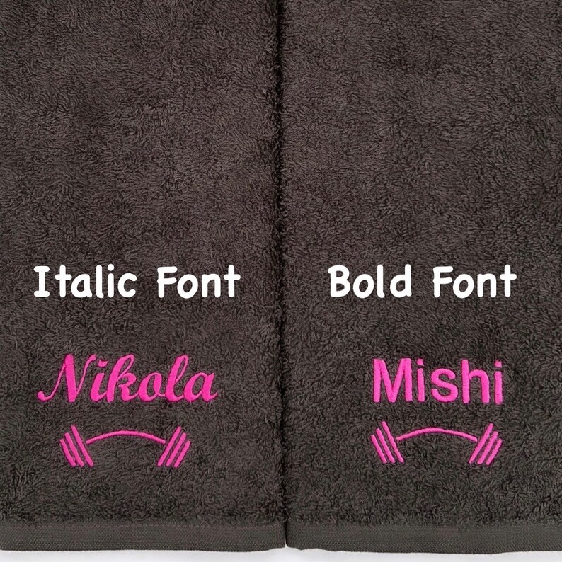 May include: Two dark gray towels with pink embroidery. The left towel has the name "Nikola" in italic font and a pink dumbbell symbol. The right towel has the name "Mishi" in bold font and a pink dumbbell symbol.