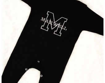 May include: Black long-sleeve baby onesie with a white embroidered letter "M" and the name "MAXWELL" underneath.