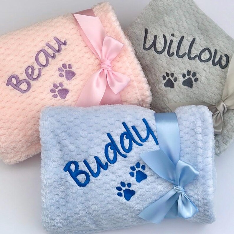 Op de afbeelding: Drie gepersonaliseerde huisdierdekens met geborduurde pootjes. De dekens zijn roze, blauw en grijs met bijpassende strikken. De namen "Beau", "Buddy" en "Willow" zijn op de dekens geborduurd.