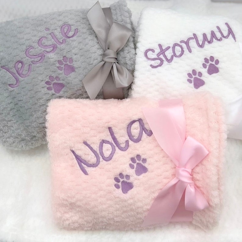 Op de afbeelding: Drie gepersonaliseerde pluche dekens met pootjes en namen geborduurd in paars. De dekens zijn grijs, wit en roze met bijpassende satijnen strikken.