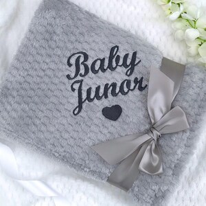 Może przedstawiać: Szary pluszowy kocyk dla dziecka z satynową wstążką zawiązaną w kokardkę. Kocyk jest haftowany tekstem "Baby Junior" i małym serduszkiem.
