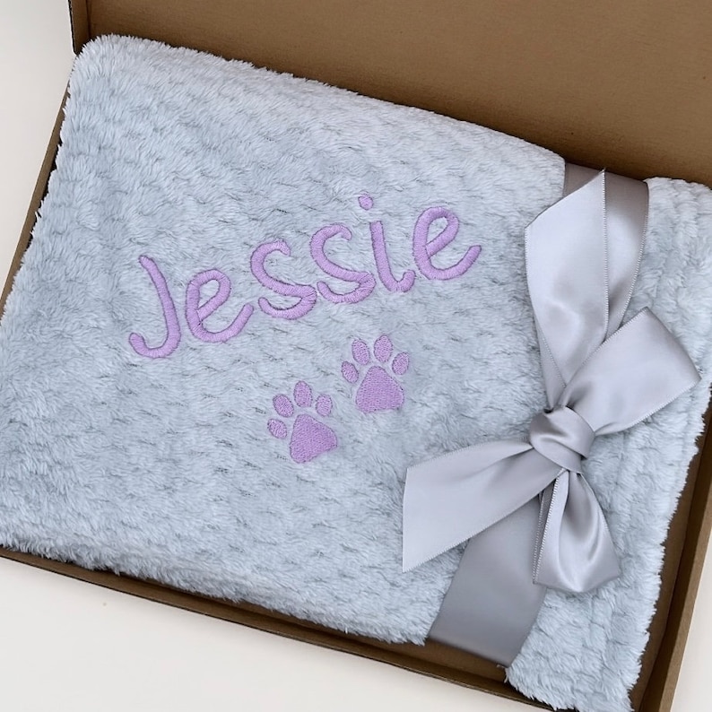 Op de afbeelding: Een lichtblauwe pluche deken met een zilveren lint dat in een strik is gebonden. De deken is gepersonaliseerd met de naam "Jessie" en twee paarse poot afdrukken.