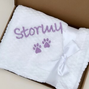 Op de afbeelding: Een wit pluche deken met een paarse geborduurde naam "Stormy" en twee poot afdrukken. Het deken is dubbelgevouwen en vastgebonden met een wit satijnen lint.