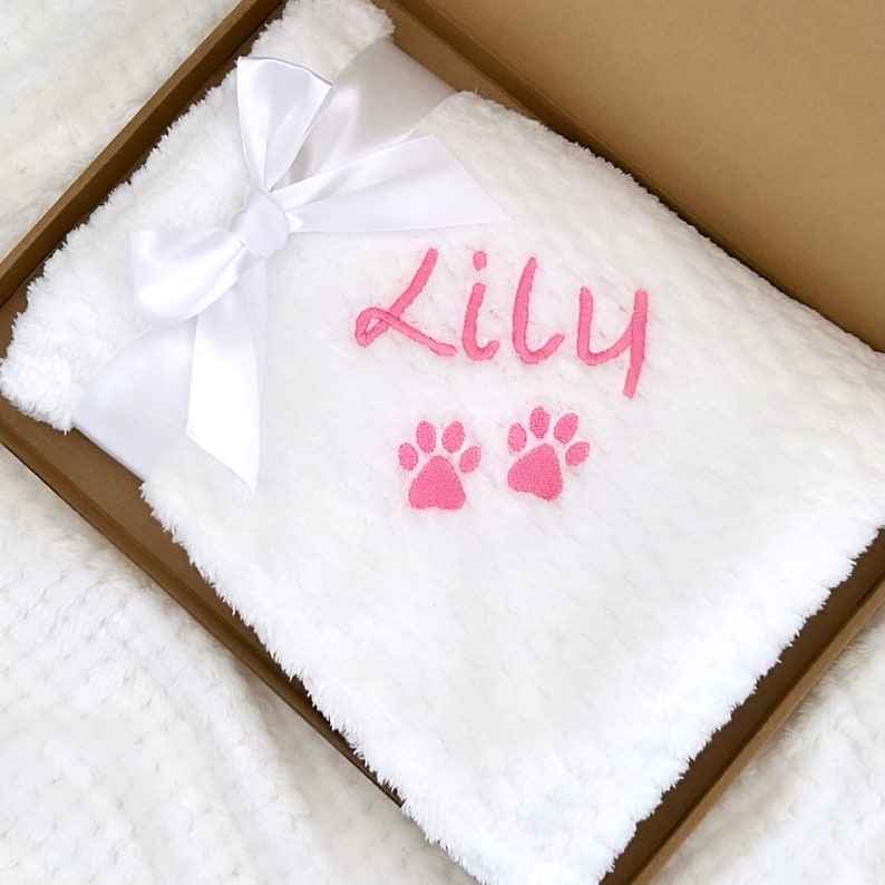 Op de afbeelding: Witte pluche deken met een witte satijnen strik. De deken is gepersonaliseerd met de naam "Lily" en twee roze poot afdrukken.