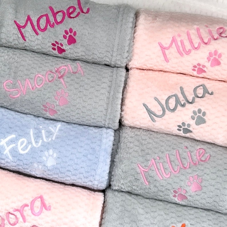 Op de afbeelding: Een stapel gepersonaliseerde grijze en roze fleece dekens met geborduurde namen en poot afdrukken. De namen zijn Mabel, Snoopy, Felix, Millie, Walla en Nora.