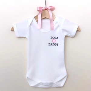 Puede incluir: Un body blanco para bebé con mangas cortas y broches rosas. El body tiene el texto "Lola" con un corazón rosa y "Daddy" bordado en la parte delantera.