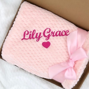 Może przedstawiać: R&oacute;żowy pluszowy koc z r&oacute;żową satynową wstążką zawiązaną w kokardę. Koc jest haftowany imieniem "Lily Grace" i małym r&oacute;żowym sercem.