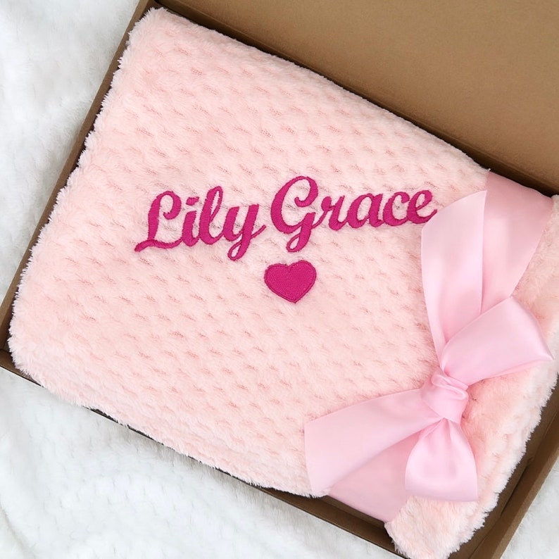 Może przedstawiać: R&oacute;żowy pluszowy koc z r&oacute;żową satynową wstążką zawiązaną w kokardę. Koc jest haftowany imieniem "Lily Grace" i małym r&oacute;żowym sercem.