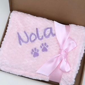 Op de afbeelding: Een roze pluche deken met een paarse geborduurde naam "Nola" en twee poot afdrukken. De deken is vastgebonden met een roze satijnen lint.