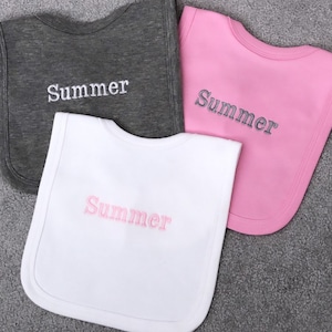 May include: Three baby bibs with the name "Summer" embroidered on them. The bibs are in grey, pink, and white.
