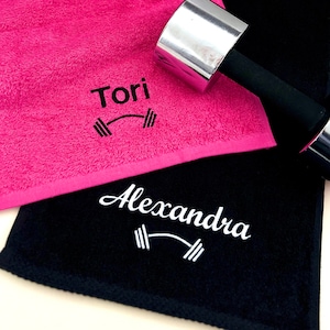 May include: Two personalized gym towels, one pink and one black, with the names "Tori" and "Alexandra" embroidered on them. Each towel features a small embroidered dumbbell graphic.