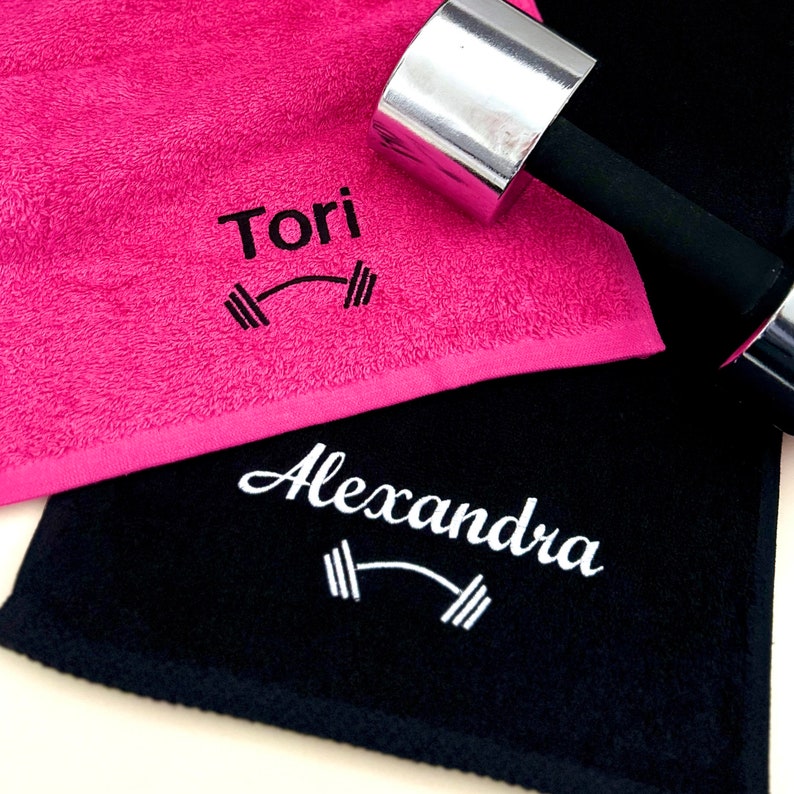 May include: Two personalized gym towels, one pink and one black, with the names "Tori" and "Alexandra" embroidered on them. Each towel features a small embroidered dumbbell graphic.