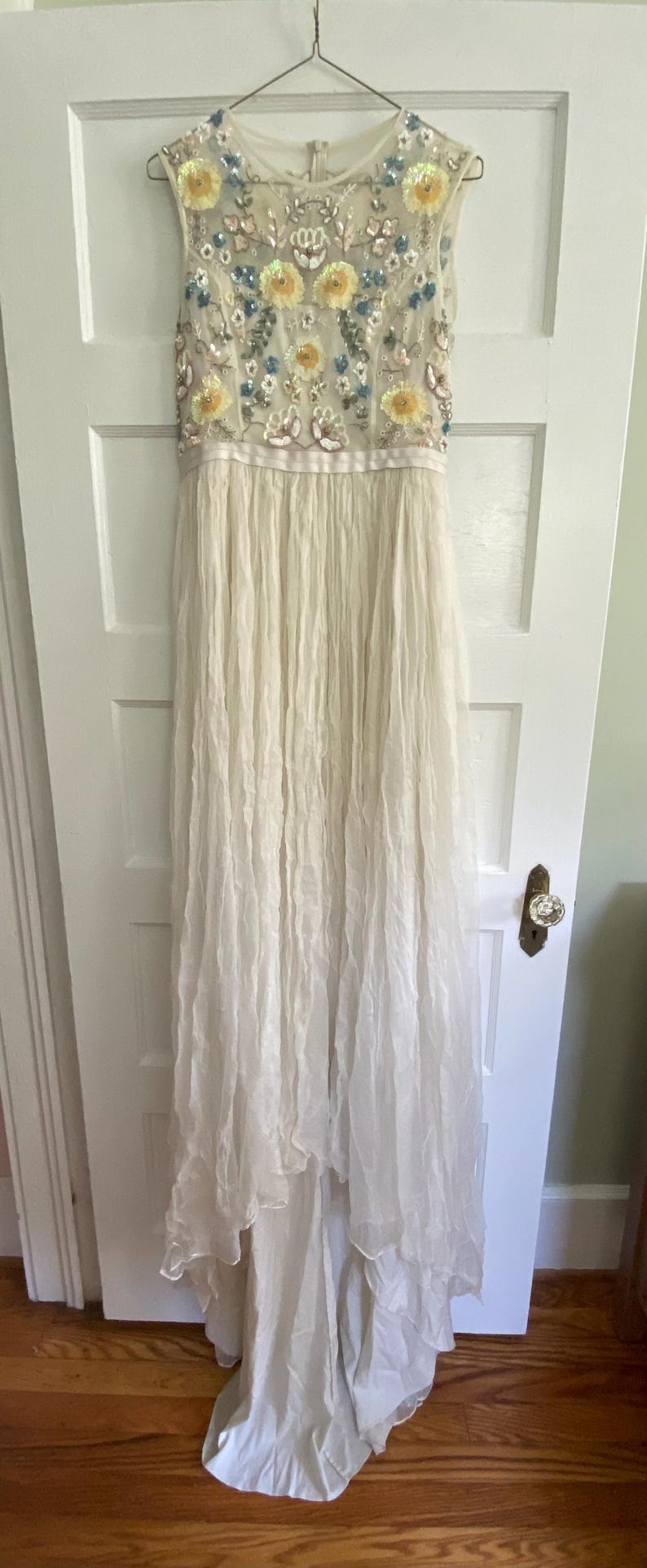 May include: A sleeveless ivory gown with a heavily embellished bodice featuring yellow and blue floral designs. The dress has a flowing, layered skirt and is hanging on a metal hanger against a white door.