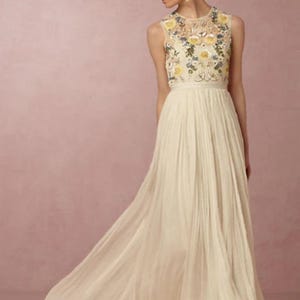 May include: A sleeveless ivory wedding dress with a floral embroidered bodice and a flowing pleated skirt. The dress features a high neckline and a long train. The floral embroidery includes yellow, blue, and green accents.