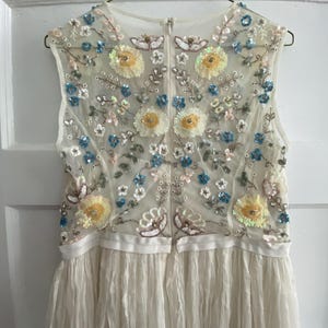 May include: Ivory sleeveless dress with floral embroidery. The bodice features a sheer overlay adorned with yellow, blue, and pink sequined flowers and leaves. The skirt is made of a pleated, flowing fabric, creating a delicate and elegant look.