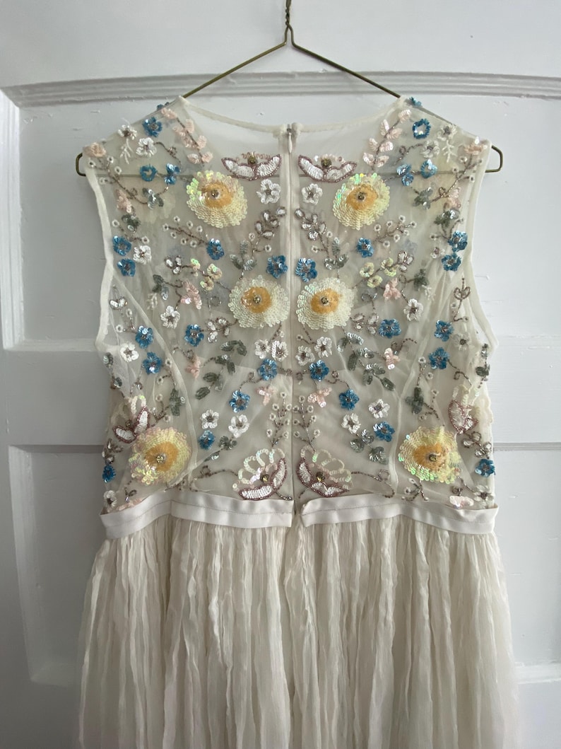 May include: Ivory sleeveless dress with floral embroidery. The bodice features a sheer overlay adorned with yellow, blue, and pink sequined flowers and leaves. The skirt is made of a pleated, flowing fabric, creating a delicate and elegant look.