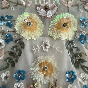 May include: Close-up of a cream-colored fabric adorned with intricate floral embroidery. The design features large, sequined flowers in shades of yellow and white, with smaller blue and white blossoms. Delicate leaves and stems add to the detailed pattern.