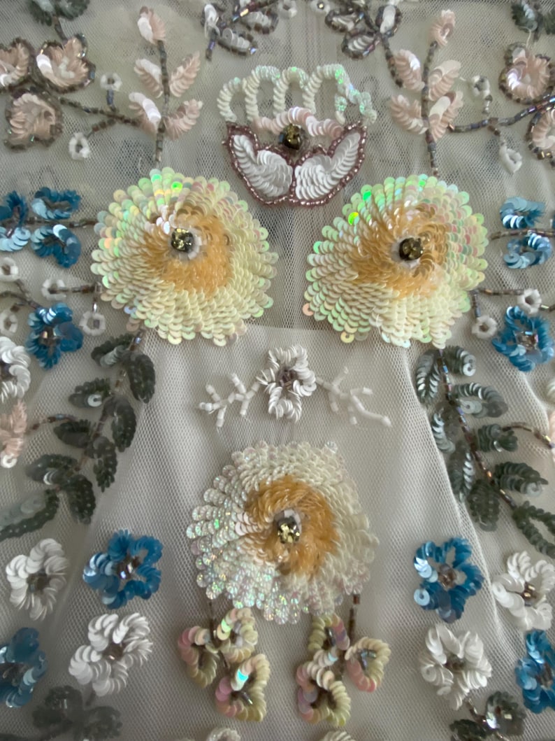 May include: Close-up of a cream-colored fabric adorned with intricate floral embroidery. The design features large, sequined flowers in shades of yellow and white, with smaller blue and white blossoms. Delicate leaves and stems add to the detailed pattern.