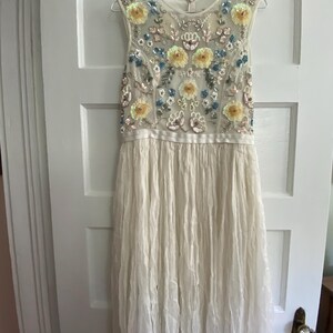May include: A sleeveless ivory dress with a floral beaded bodice. The bodice features yellow, blue, and silver floral embellishments. The skirt is a flowing, pleated fabric. The dress hangs on a hanger against a white door.