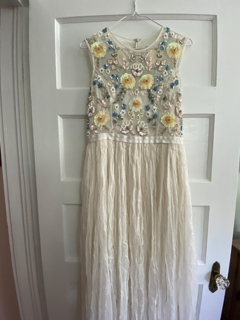 May include: A sleeveless ivory dress with a floral beaded bodice. The bodice features yellow, blue, and silver floral embellishments. The skirt is a flowing, pleated fabric. The dress hangs on a hanger against a white door.