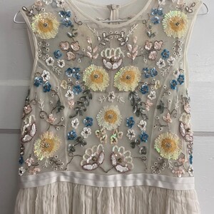 May include: A sleeveless ivory dress with floral embroidery. The bodice features a sheer overlay adorned with yellow, blue, and pink sequined flowers. The skirt is pleated, and the dress hangs on a silver hanger.