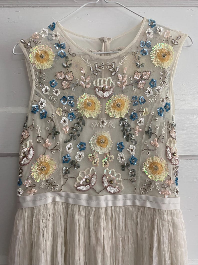 May include: A sleeveless ivory dress with floral embroidery. The bodice features a sheer overlay adorned with yellow, blue, and pink sequined flowers. The skirt is pleated, and the dress hangs on a silver hanger.