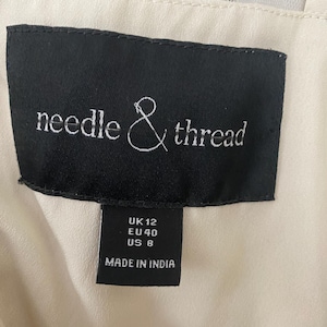 May include: A close-up of a black label with the words "needle & thread" in white script. Below the brand name are size indicators: UK 12, EU 40, US 8. The label reads "MADE IN INDIA". The background is a cream-colored fabric.