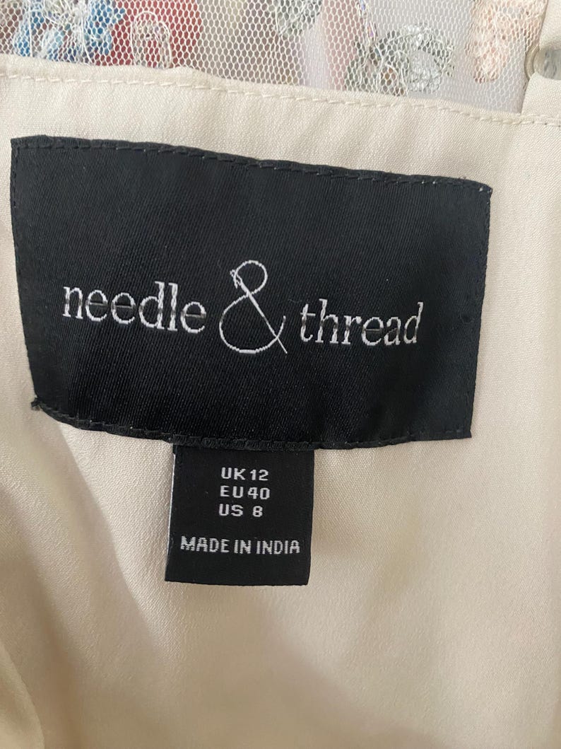 May include: A close-up of a black label with the words "needle & thread" in white script. Below the brand name are size indicators: UK 12, EU 40, US 8. The label reads "MADE IN INDIA". The background is a cream-colored fabric.