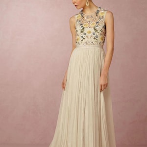 May include: A sleeveless ivory gown with a floral embroidered bodice. The dress features a pleated skirt that flows to the floor. The bodice is adorned with yellow and blue floral embroidery. The model is wearing gold earrings and has her hair up.