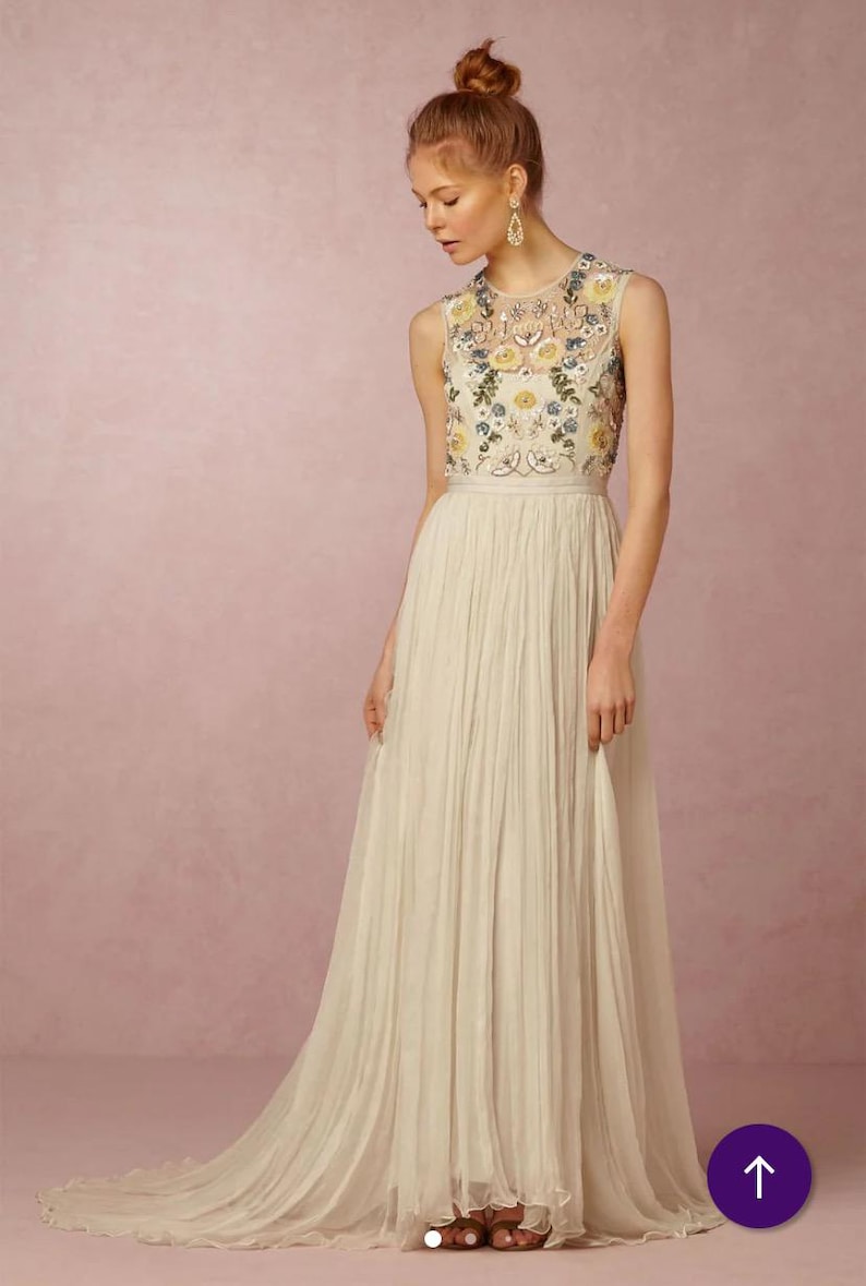 May include: A sleeveless ivory gown with a floral embroidered bodice. The dress features a pleated skirt that flows to the floor. The bodice is adorned with yellow and blue floral embroidery. The model is wearing gold earrings and has her hair up.