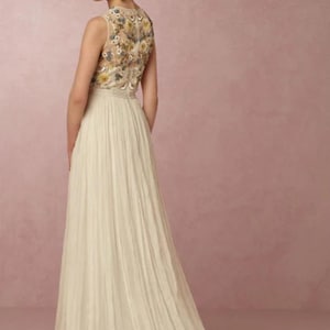 May include: A sleeveless ivory wedding gown with a floral embroidered bodice and a flowing pleated skirt. The back of the dress features a detailed floral pattern in shades of yellow, blue, and cream. The dress has a long train.