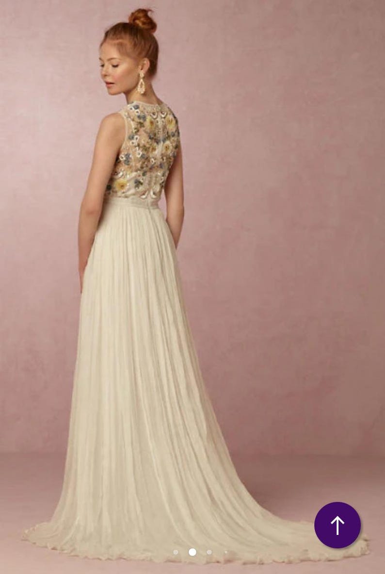 May include: A sleeveless ivory wedding gown with a floral embroidered bodice and a flowing pleated skirt. The back of the dress features a detailed floral pattern in shades of yellow, blue, and cream. The dress has a long train.