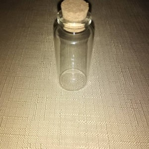 Peut inclure: Une petite fiole en verre transparent avec un corps cylindrique et une base arrondie. L'ouverture supérieure est scellée par un bouchon en liège texturé de couleur brun clair qui s'insère parfaitement dans le goulot de la bouteille.