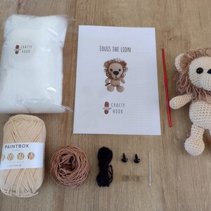 May include: A crochet lion kit with instructions, yarn, stuffing, safety eyes, and a crochet hook. The kit includes beige and brown yarn, a crochet hook, and two black safety eyes.