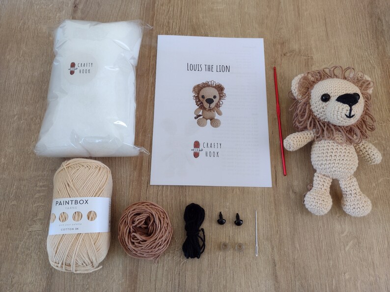 May include: A crochet lion kit with instructions, yarn, stuffing, safety eyes, and a crochet hook. The kit includes beige and brown yarn, a crochet hook, and two black safety eyes.