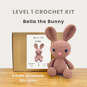 Könnte beinhalten: Ein Häkelset für Bella das Häschen, ein Projekt der Stufe 1. Das Set enthält alle Materialien, um ein rosa gehäkeltes Häschen zu erstellen. Der Text "LEVEL 1 CROCHET KIT" und "Bella the Bunny" sind oben im Bild zu sehen.
