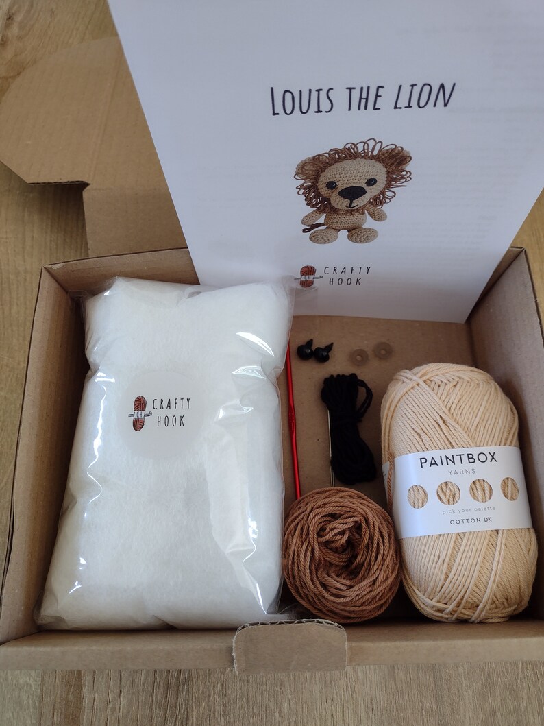 May include: A crochet kit for making a lion amigurumi toy. The kit includes beige and brown yarn, stuffing, safety eyes, a crochet hook, and instructions. The kit is packaged in a brown cardboard box with the words "Louis the Lion" and "Crafty Hook" printed on the lid.
