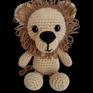 May include: A crocheted lion toy with a brown mane, black eyes, and a black nose. The lion is sitting with its legs crossed and has a brown tail.