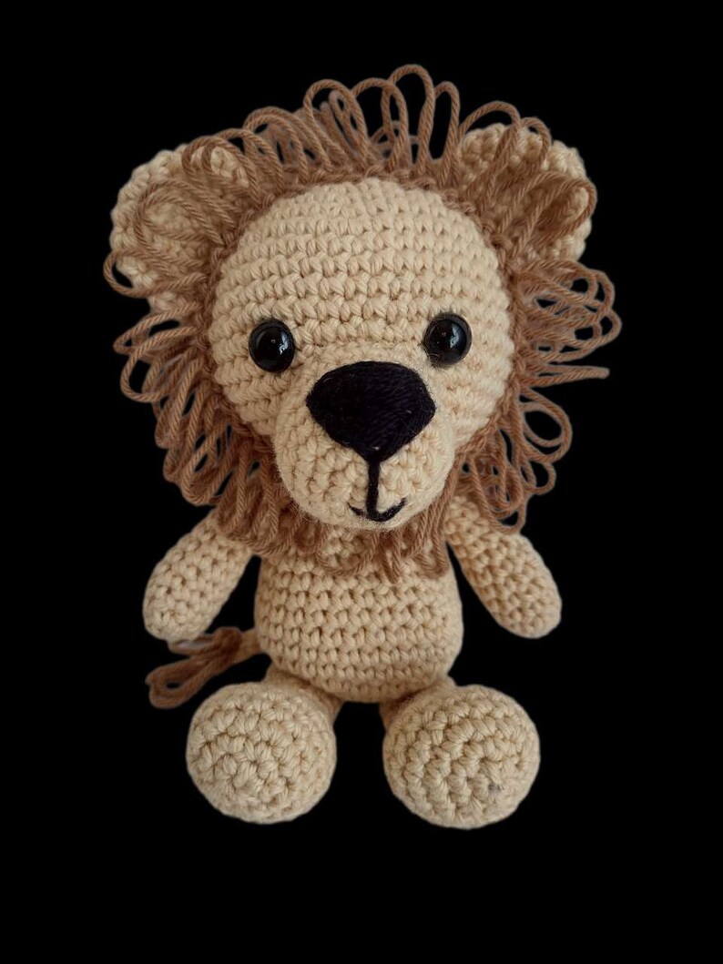 May include: A crocheted lion toy with a brown mane, black eyes, and a black nose. The lion is sitting with its legs crossed and has a brown tail.