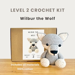 Könnte beinhalten: Ein Häkelset der Stufe 2 mit einem grau-weißen gehäkelten Wolf namens Wilbur. Das Set enthält alle Materialien und besteht aus 100 % Baumwolle. Auf der Box steht "LEVEL 2 CROCHET KIT" und "Wilbur the Wolf".