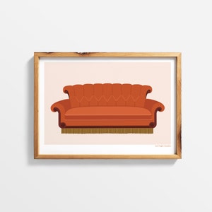 May include: A print of a brown sofa with a tufted back and fringe trim. The sofa is set against a light pink background. The print is framed in a light brown wood frame. The text "by Faegi Studios" is in the bottom right corner of the print.
