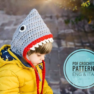 Può includere: Un cappello a forma di squalo in crochet grigio con denti rossi e bianchi. Il cappello è indossato da un bambino. L'immagine include il testo "PDF CROCHET PATTERN ENG & ITA".