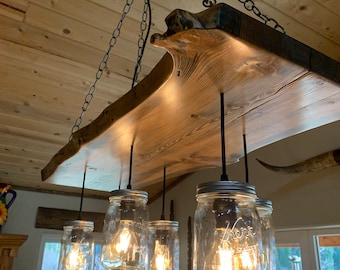 May include: A rustic wooden chandelier with six mason jar lights hanging from it. The chandelier is made from a thick slab of wood with a natural edge. The mason jars are clear glass and have silver lids. The lights are turned on and are illuminating the room.