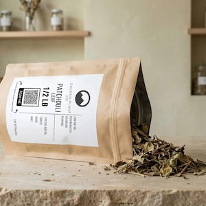 May include: A tan stand-up pouch containing dried patchouli leaves, with a white label reading Sacred Plant Co, Patchouli Leaf, 1/2 LB, and a QR code. The pouch is tilted, spilling some of the brown, textured dried leaves onto the surface.
