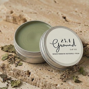 May include: An open, silver-colored tin of green balm with the text "25.1 Ground" and "Estate Reserve: Patchouli + Tulsi" on the lid. The tin sits on a textured, light brown surface with dried leaves.
