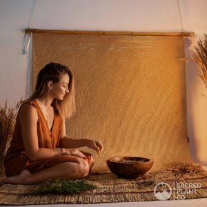 Woman seated on a woven mat in a warm, rustic setting, handling premium organic bulk oatstraw herb in a wooden bowl, surrounded by dried grasses and green oats, highlighting the natural wellness of Avena Sativa with Sacred Plant Co branding.