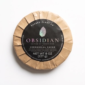 Könnte beinhalten: Eine kreisförmige Packung Obsidian zeremonieller Kakao, eingewickelt in braunes Papier mit einem schwarzen Etikett. Das Etikett zeigt ein geometrisches Kristallsymbol, den Text Sacred Plant Co, Obsidian, Edition of Twenty-Five und 227 g.