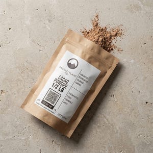 May include: A brown paper pouch of Sacred Plant Co. Cacao Powder 1/2 LB with a white label featuring a mountain logo, a QR code, and text detailing low water regenerative Colorado farming, plus tasting notes of citrus, chamomile, and earth.