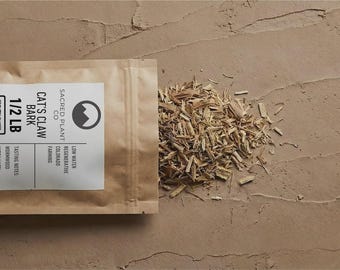 May include: A brown paper pouch labelled Cat's Claw Bark 1/2 LB with a QR code and Sacred Plant Co logo, containing dried, light-coloured woody bark pieces. The pouch is open, with a portion of the shredded bark spilled out onto the surface.