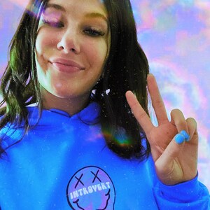 May include: A person wearing a bright blue hoodie with the word "INTROVERT" inside a melting smiley face graphic. The person is giving a peace sign with blue painted nails. The background has a colorful, dreamy effect.